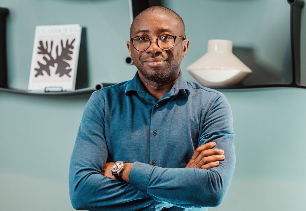An image of a man named Ola Oyetayo who is the founder of a company called Verto, a winner of the Milken-Motsepe Prize. He has a bald head, brown skin, and a slight beard and is wearing round glasses, a blue collared shirt, and standing in front of a blue decorative background with crossed arms and slight smile.