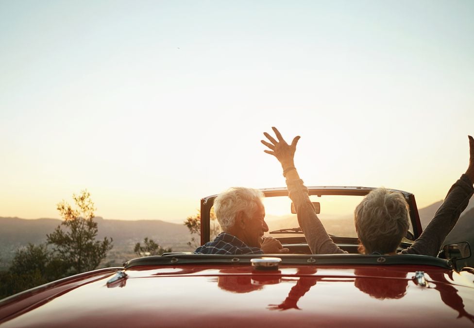 An image of two people's silhouettes in a red convertible, driving into the sunset with one of them holding his or her hands in the air.