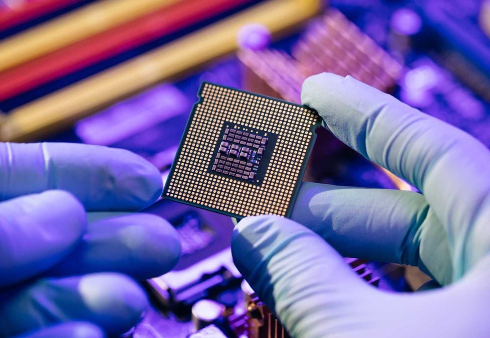 An image of hands with light purple gloves holding what appears to be a computer chip with a purple-ish background of technical computer pieces.