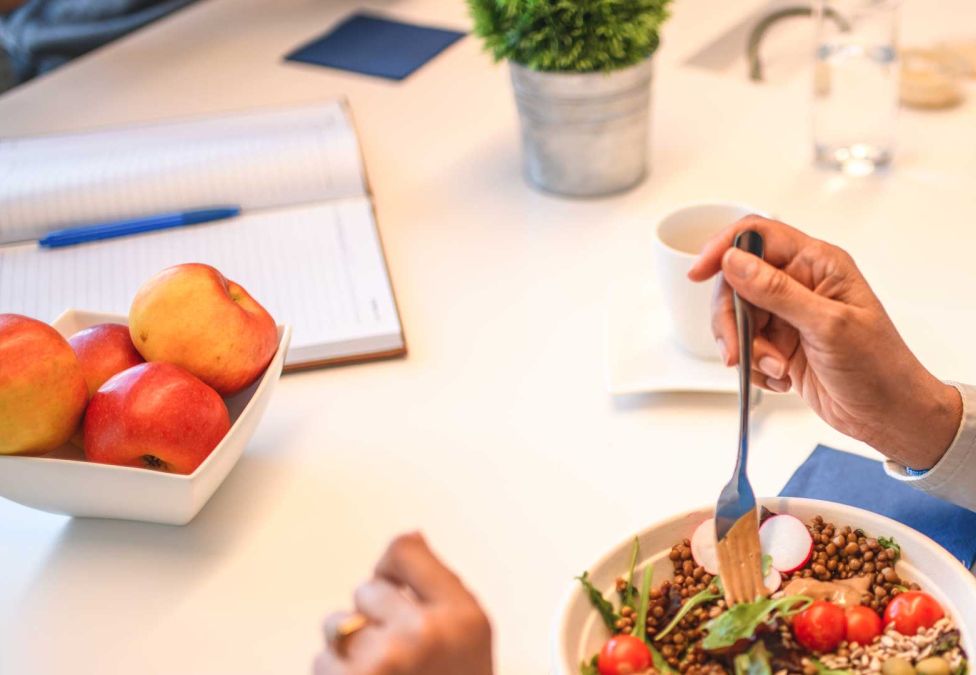 An image of someone's hands eating a bowl of food with various vegetables, and nearby sits a white bowl full of red apples.