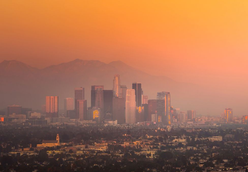 A wide view of Los Angeles with dark smoke billowing from wildfires in the distance, rising behind downtown buildings and palm trees. The sky is streaked with orange and gray, emphasizing the scale of the disaster. The image reflects the urgency and impact of wildfire recovery efforts in the city.
