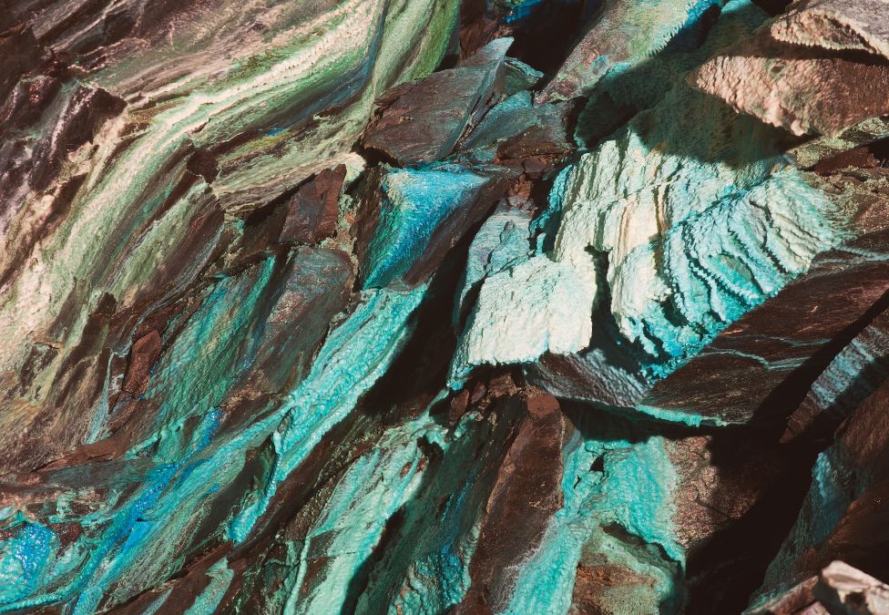 Close-up of jagged rock layers with shimmering turquoise, teal, and brown mineral streaks creating a textured, iridescent surface.