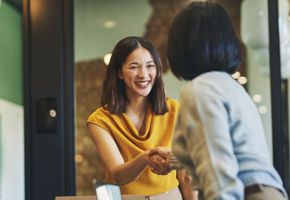 Two people in a professional setting smiling and shaking hands, symbolizing agreement or partnership.