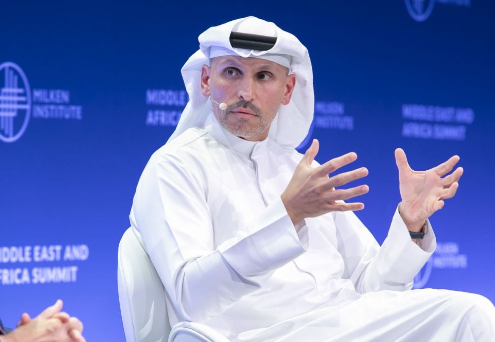 A man in traditional white Middle Eastern attire speaks on stage at the Milken Institute Middle East and Africa Summit. He gestures with both hands as he addresses the audience, wearing a headset microphone against a blue backdrop.