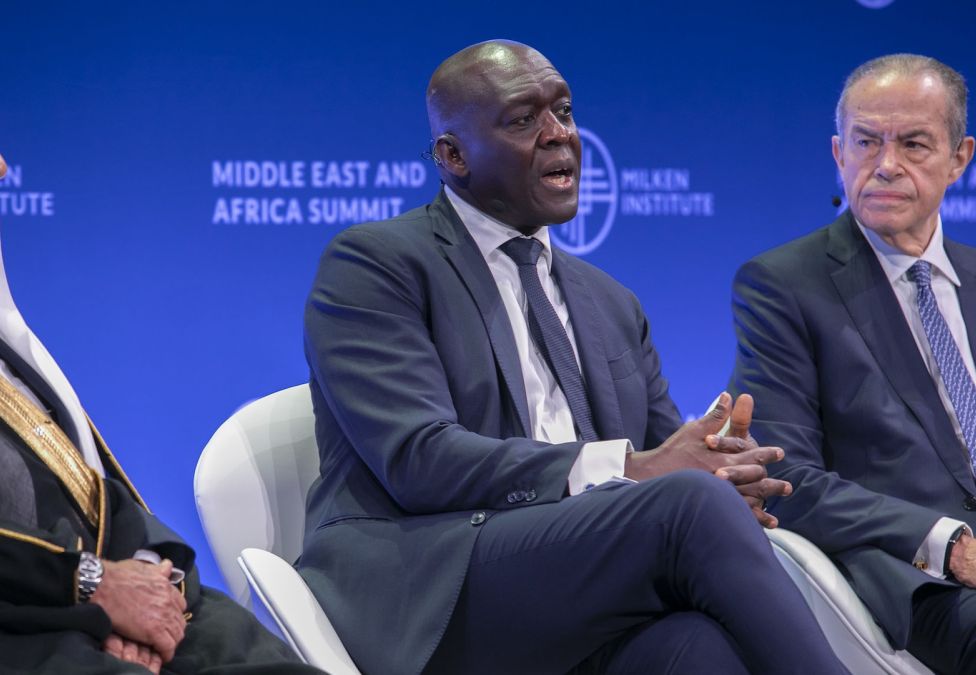Three men seated on stage during a panel at the Milken Institute Middle East and Africa Summit. The man in the center, wearing a dark suit and tie, is speaking with his hands clasped. On his left, a man in traditional Middle Eastern attire listens, while on his right, another man in a suit looks on attentively.