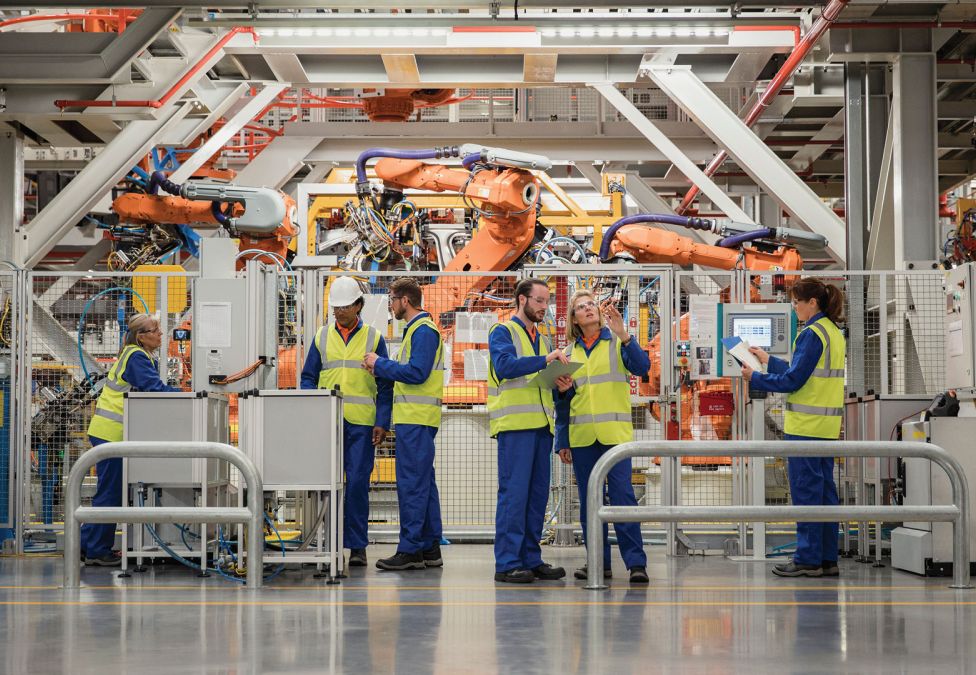 Workers in high-visibility vests and hard hats operate and monitor machinery in a modern factory with large robotic arms.