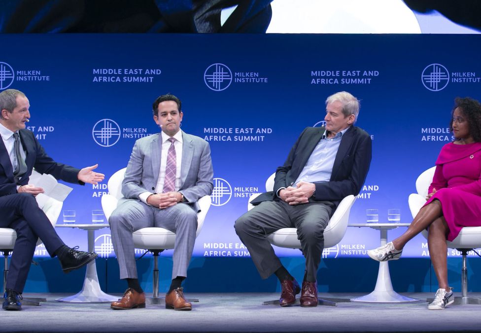 Four panelists seated on stage at the Milken Institute Middle East and Africa Summit. A moderator on the left gestures while holding notes, speaking to three panelists: a man in a gray suit, a man in a dark blazer and open-collared shirt, and a woman in a bright pink dress with sneakers. The backdrop features the Milken Institute logo and event name.