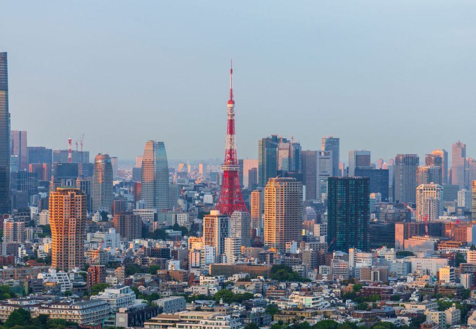 A panoramic view of Tokyo’s skyline at sunset, featuring the iconic red and white Tokyo Tower in the center, surrounded by modern high-rise buildings and skyscrapers with golden light reflecting off their glass facades.