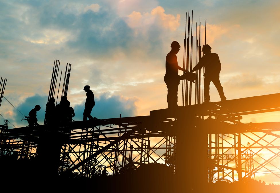 Silhouetted construction workers on elevated scaffolding at sunset, adjusting rebar and walking across metal framework against an orange-blue sky.