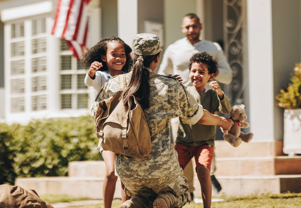 A service member in uniform kneels to embrace two excited children in front of their home.