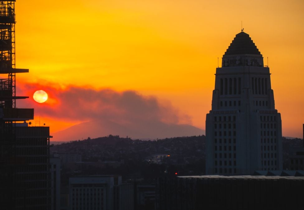 Los Angeles City Hall silhouetted at sunset as wildfire smoke drifts across the skyline, with an orange sky and the sun partially obscured.