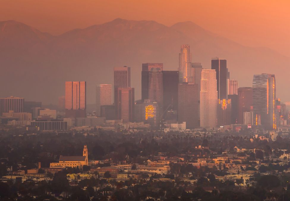 Los Angeles skyline at sunset with downtown buildings silhouetted against hazy mountains.