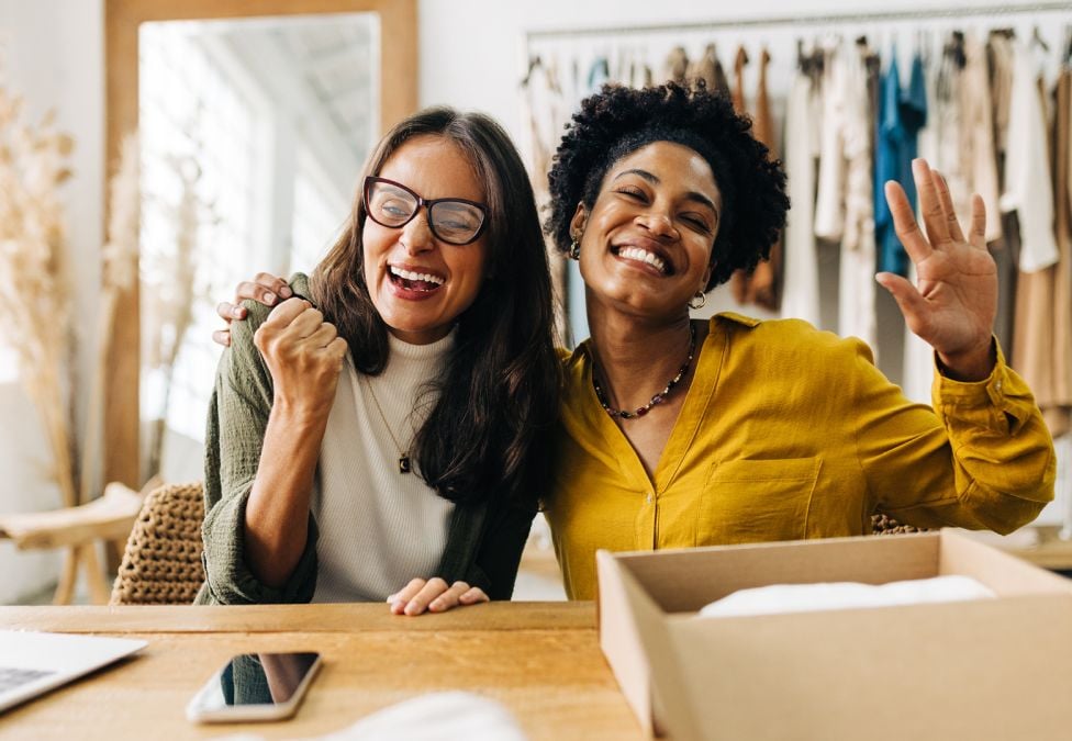 Two women smiling and waving while seated at a table in a studio, with clothing racks visible in the background.