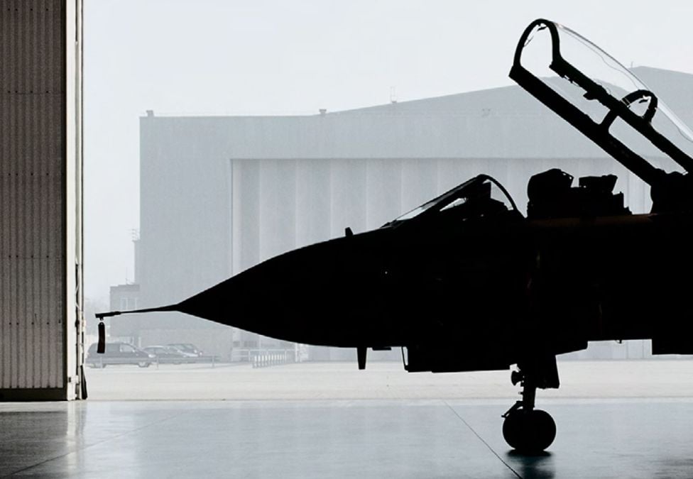 A dramatic silhouette of a fighter jet parked inside a hangar, with the cockpit canopy open and a misty airfield visible through the large hangar doors.