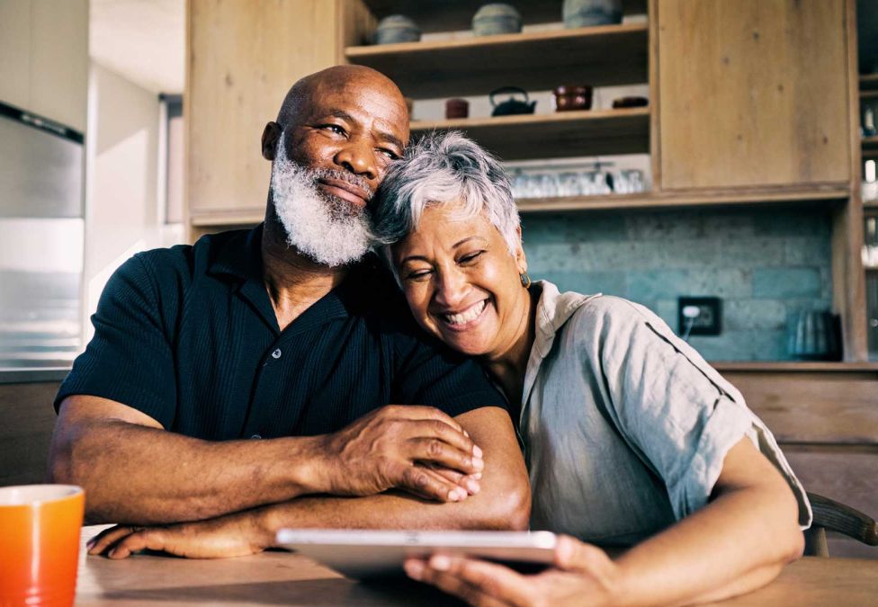 A smiling older Black man with a white beard and a woman with short grey hair share an affectionate moment in a kitchen.