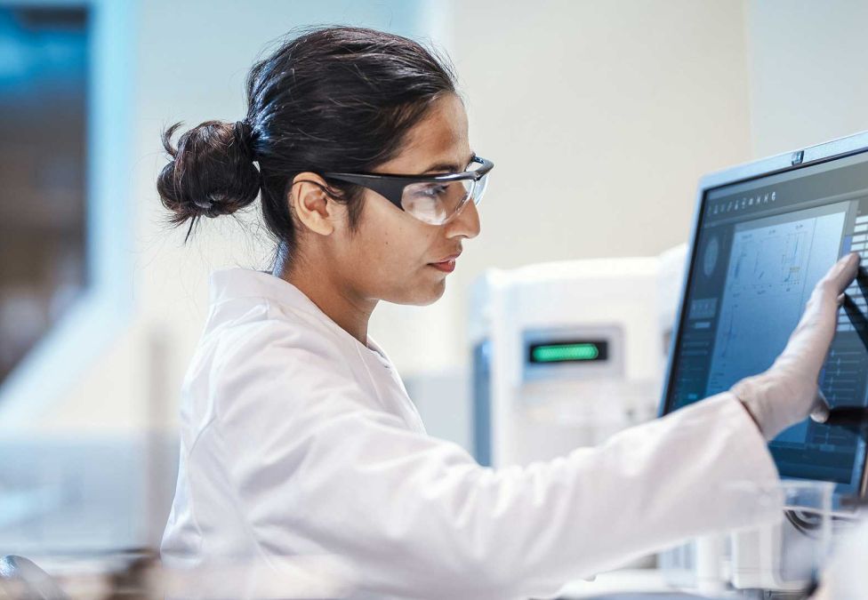 A female scientist in a white lab coat and safety goggles focuses intently while interacting with a touch-screen monitor in a modern laboratory setting.