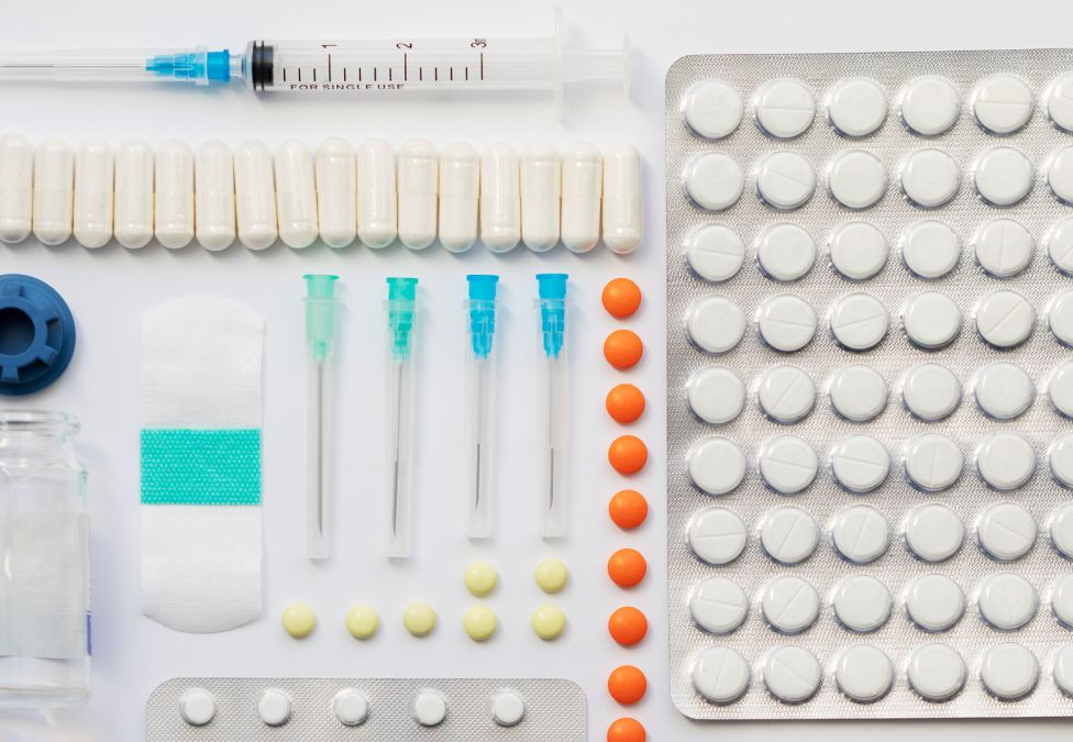 A high-angle, organized flat lay of pharmaceutical supplies on a white background, including a syringe, a large blister pack of white pills, a row of capsules, sterile needles, and loose orange tablets.