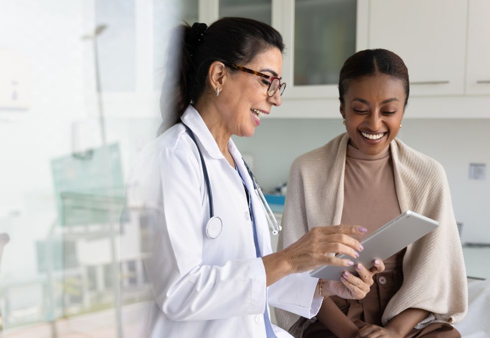 A doctor in a white coat and stethoscope smiles while showing a tablet to a patient seated on an exam table in a medical office. Both women are smiling and engaged with the screen.