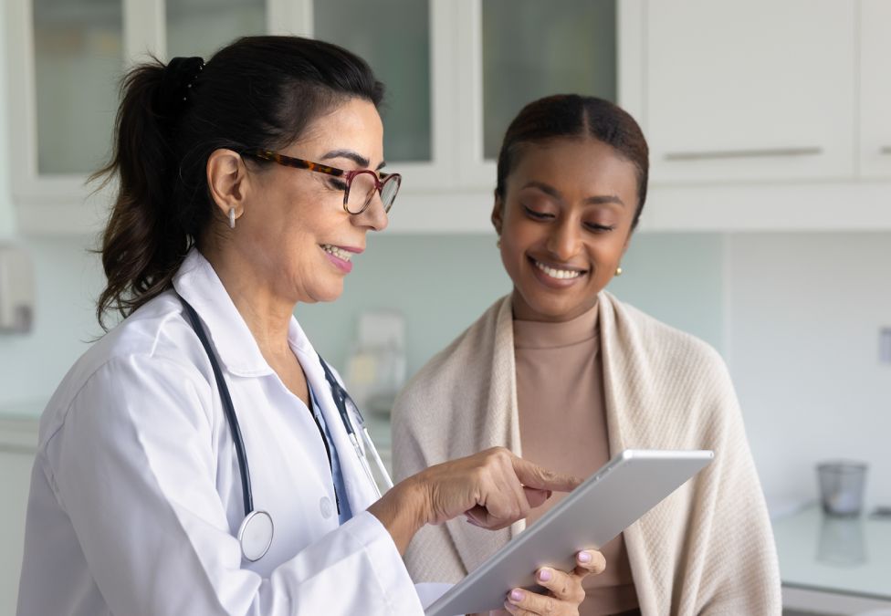 A female doctor in a white coat and stethoscope shows information on a tablet to a smiling female patient during a medical consultation.