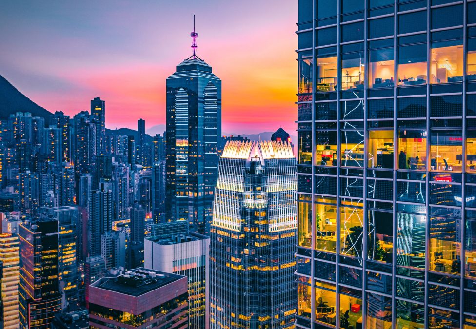 Modern glass office towers in Hong Kong at dusk with illuminated interiors and surrounding urban skyline against a colorful sunset.