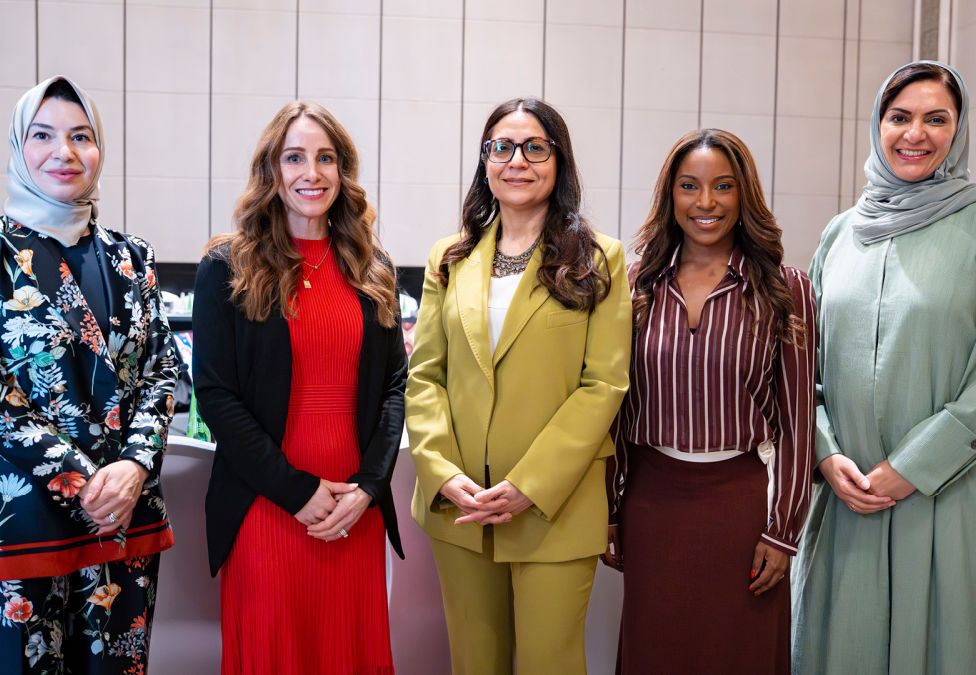 Five professionally dressed women from diverse backgrounds stand together smiling in a conference room.