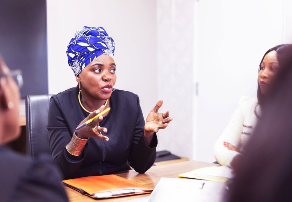 A woman in a blue patterned head wrap speaks animatedly during a business meeting with colleagues.