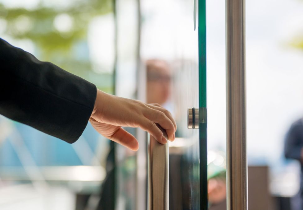A hand in a black sleeve reaching to open a glass office building door with a stainless steel handle.
