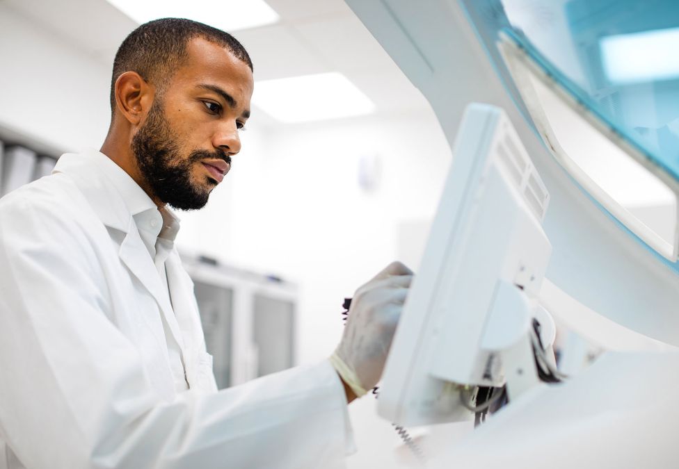 A scientist in a white lab coat and gloves operating medical or laboratory equipment in a bright clinical setting.