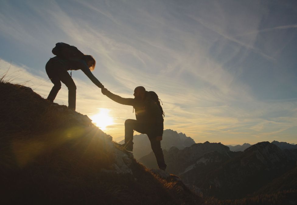 Two hikers silhouetted against a sunset, one reaching down to help the other climb a rocky mountain slope.