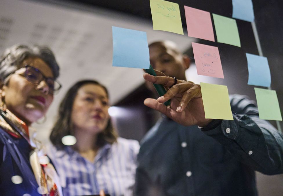 Three colleagues collaborating at a glass wall covered in colorful sticky notes during a brainstorming session.