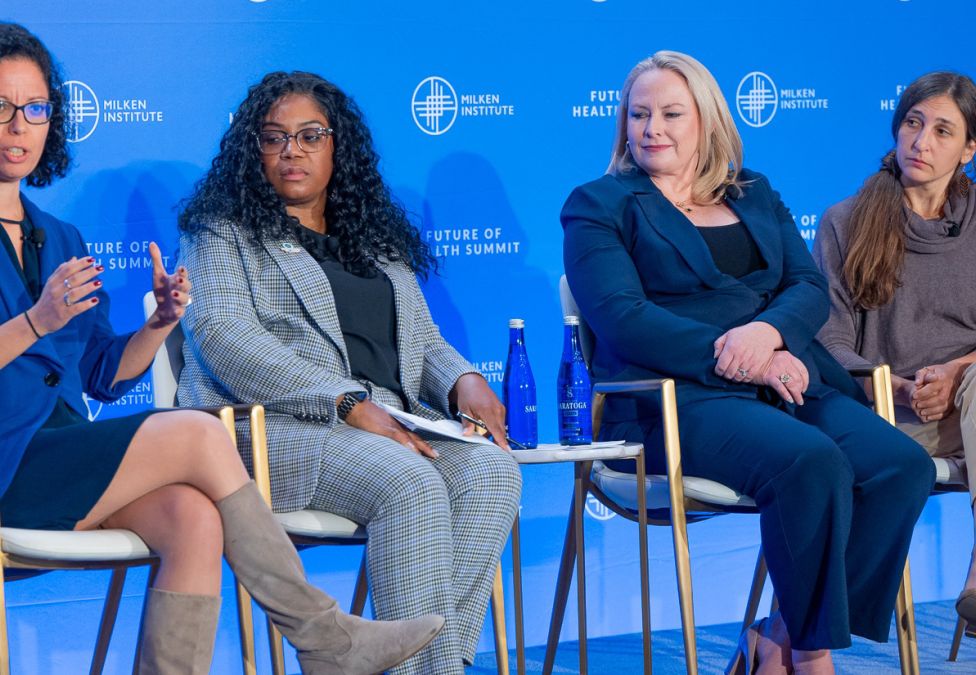 Four professional women seated on white chairs during a panel discussion against a blue backdrop