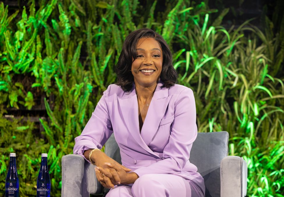 Woman in lavender suit smiling while seated outdoors with lush green foliage background