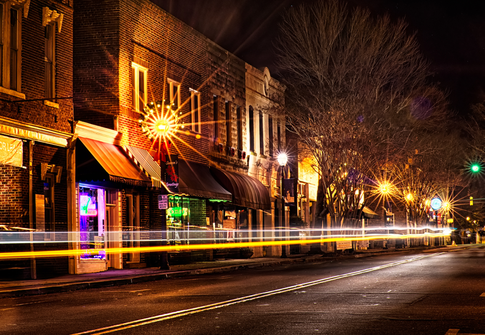 Historic downtown street at night with illuminated storefronts and light trails from passing traffic