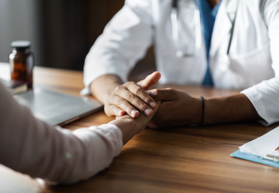 Doctor in white coat with stethoscope holding patient's hands across wooden desk