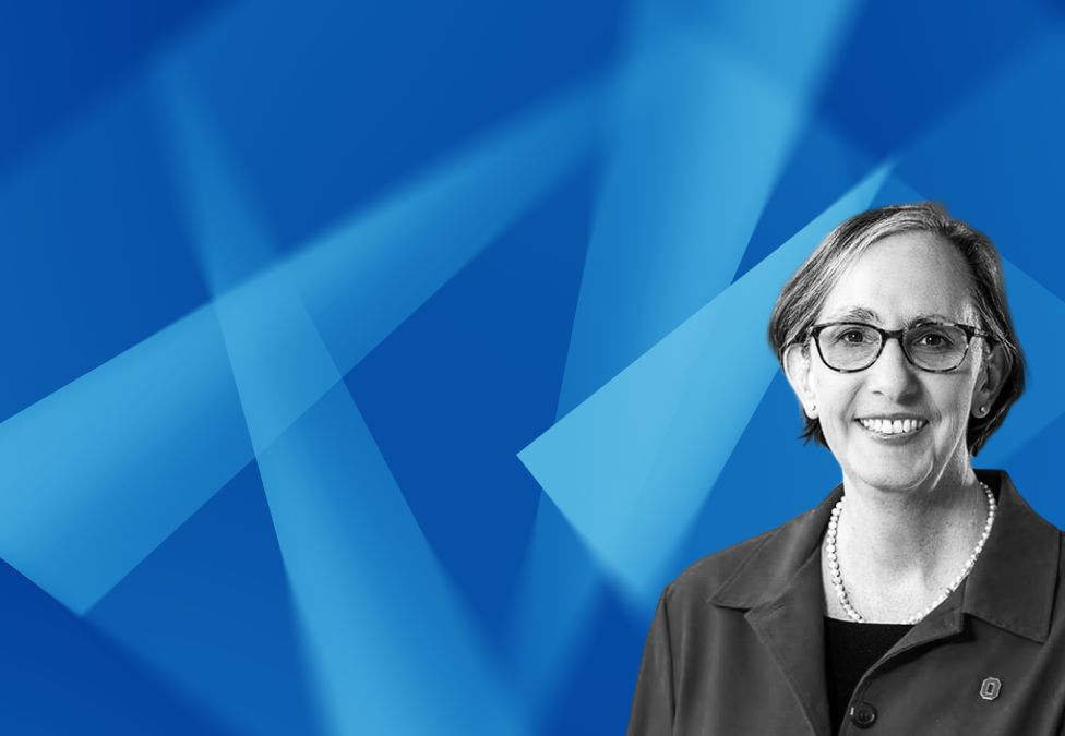 Professional headshot of a smiling woman with glasses against a blue geometric background