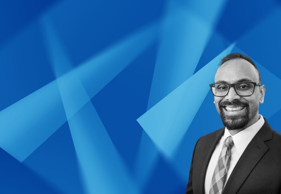 Professional headshot of smiling man in suit and glasses against blue geometric background