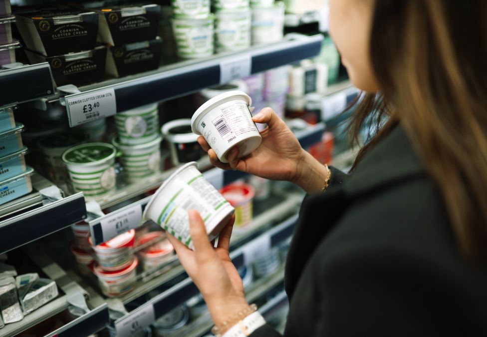 Person in dairy aisle of grocery store comparing two containers of what appears to be yogurt