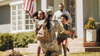 A service member in uniform kneels to embrace two excited children in front of their home.